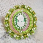 Bague Anisse brodée d’un cabochon résine représentant une femme et des roses, ornée de rocailles Vert Anis, Rose, Jaune Nacré et Ivoire. Elle est finie avec des toupies et perles sur bague réglable 